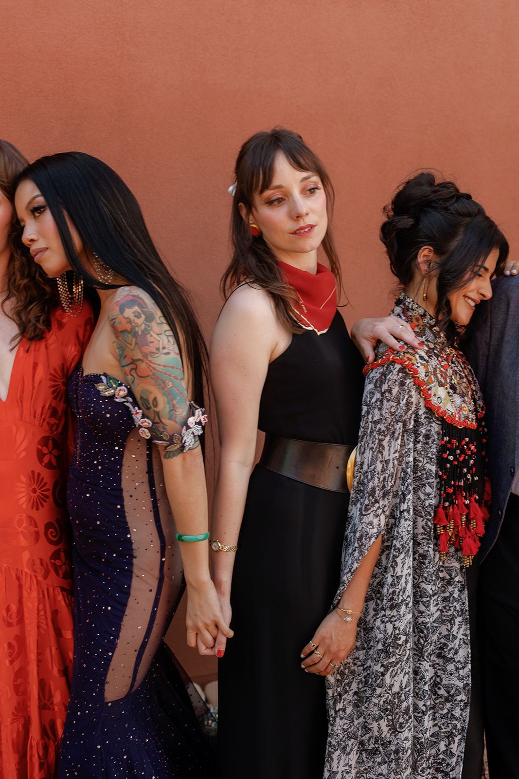 A group of women in formal evening wear including sparkly dresses and red accents pose together against a pink backdrop.
