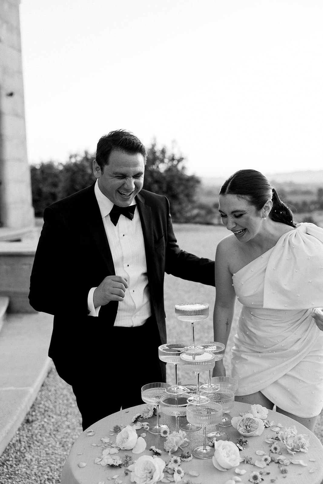 Newlywed couple sharing a joyful moment while cutting their elegant tiered wedding cake with champagne glasses.
