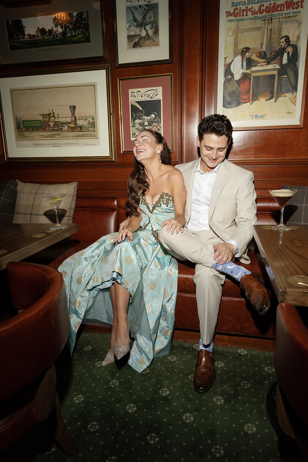 A couple shares a joyful moment at a restaurant, with decorative vintage prints on wood-paneled walls behind them.