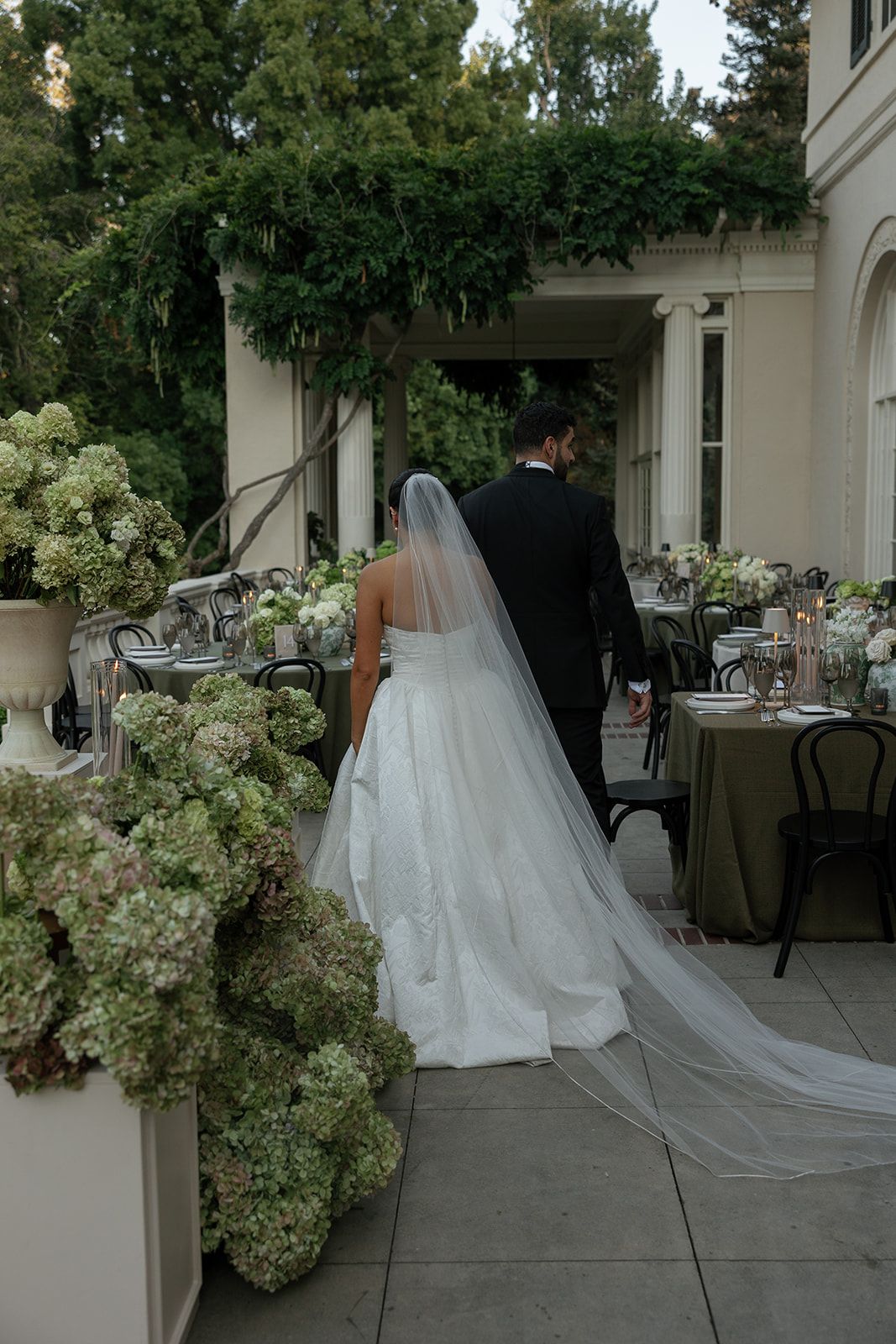 A wedding couple walks along a covered outdoor patio decorated with lush white hydrangea floral arrangements.