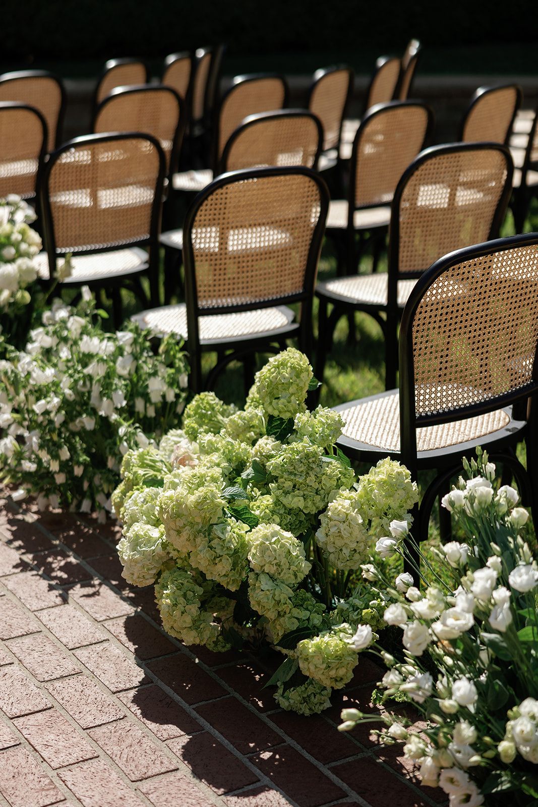 Elegant cane-back wooden chairs arranged in rows with white hydrangeas lining the aisle on a brick paver patio.