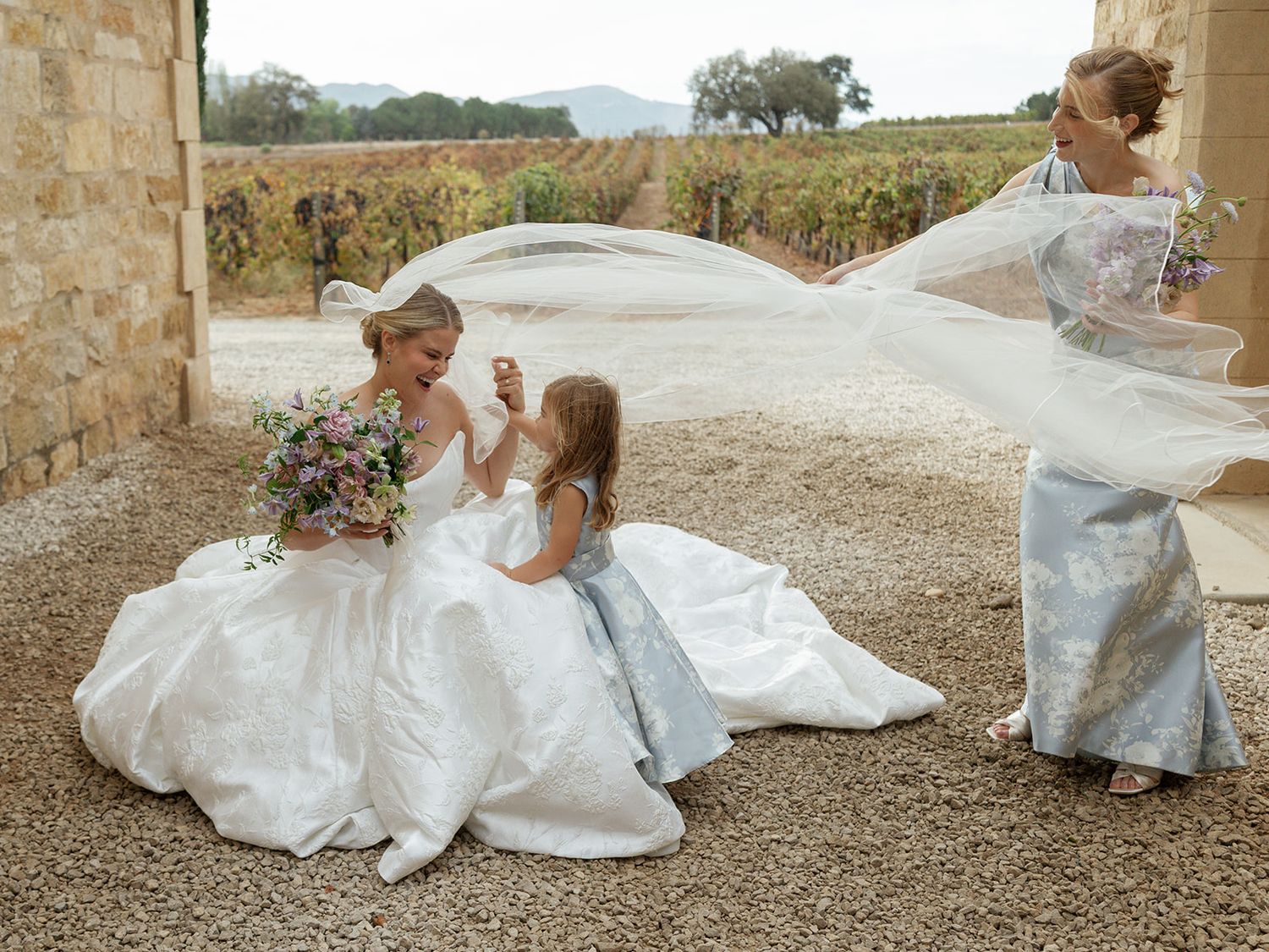 Bride in white gown sits with flower girl in blue dress outdoors at vineyard wedding venue.