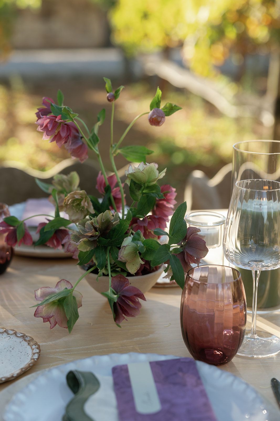 Elegant outdoor table setting with purple hellebore floral centerpiece, tinted wine glasses, and lavender linen napkins on wood table.