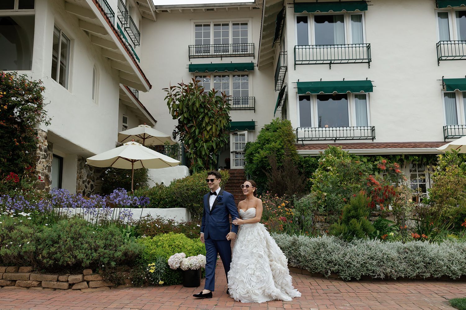 A couple stands on a garden terrace in front of a white hotel with green shutters and blooming flower beds.