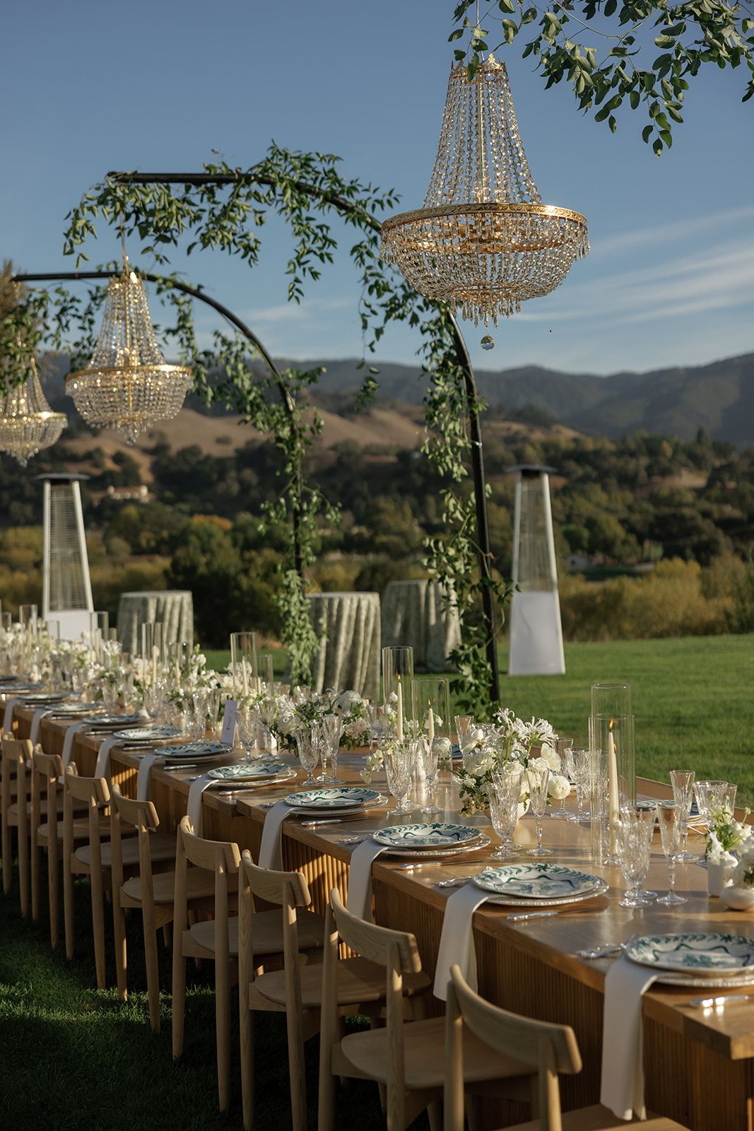 Outdoor wedding reception setup with long tables and crystal chandeliers against a mountain backdrop.