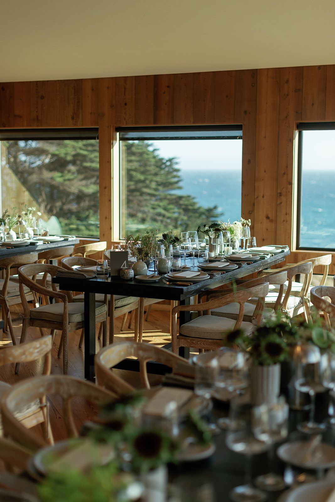Elegant coastal wedding reception venue with wooden chairs, floral centerpieces, and ocean views through large windows.