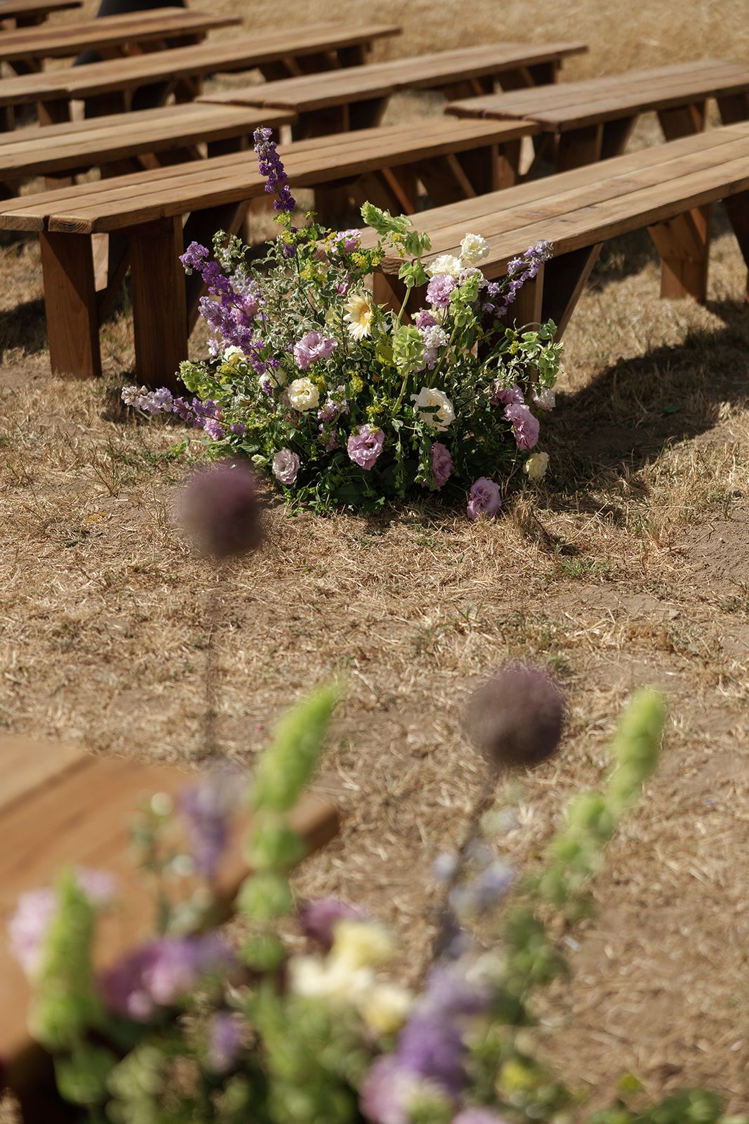 Rustic wooden benches arranged outdoors for a wedding ceremony, adorned with purple and white wildflower arrangements on dry grass.