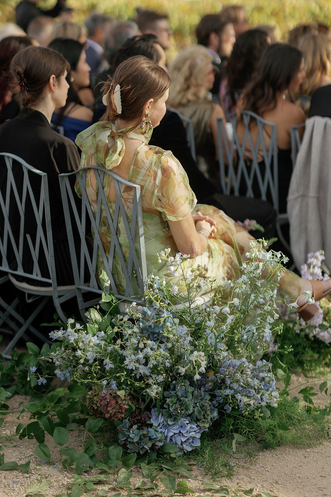 Wedding guest in yellow floral dress seated beside lush blue hydrangea and greenery ceremony aisle flowers.