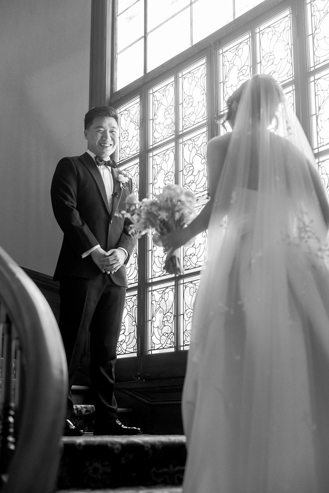 Black and white photo of a groom smiling at his bride in a wedding gown holding bouquet near stained glass windows.