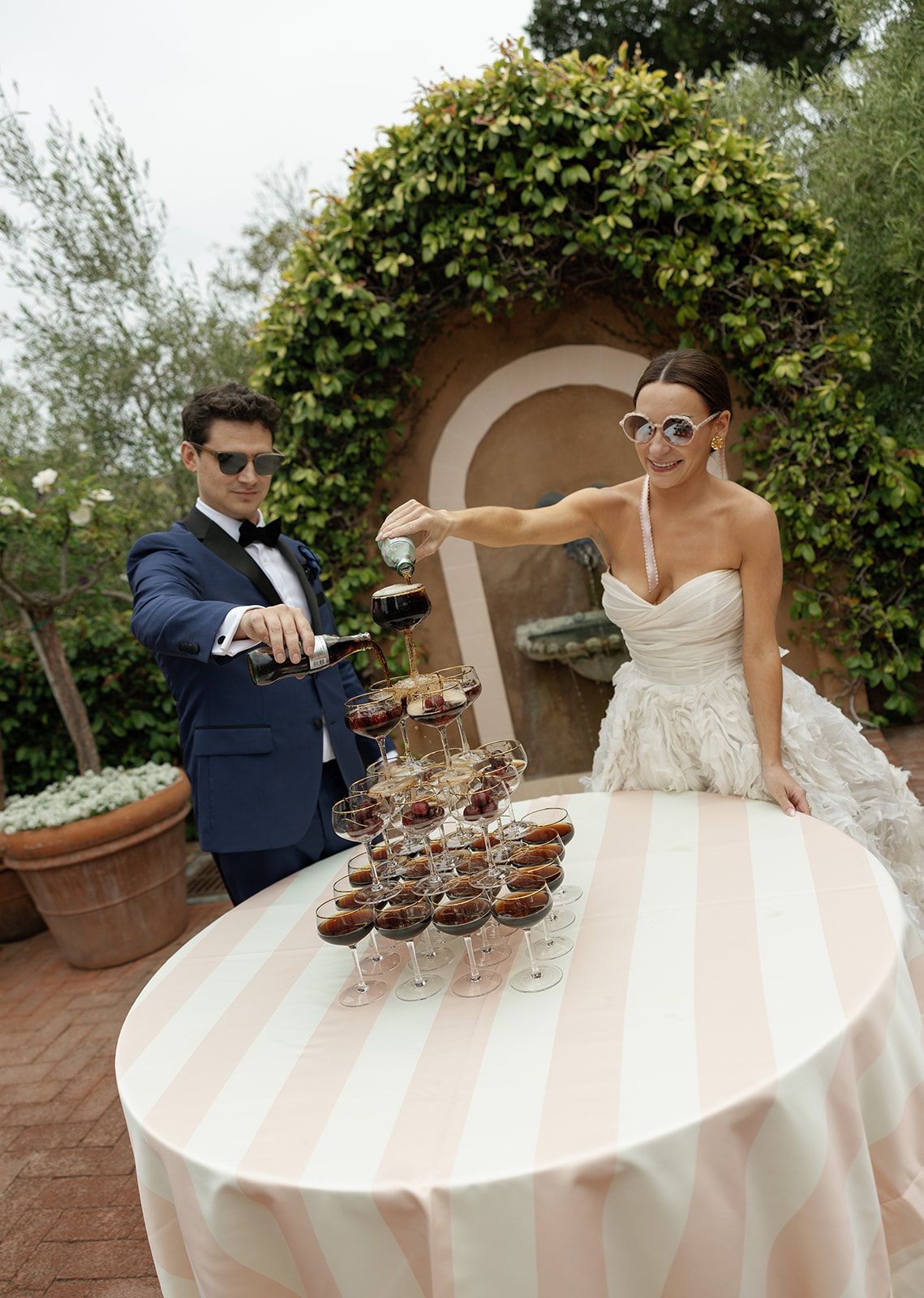 A bride and groom pour champagne together into a tower of glasses at an outdoor wedding reception.