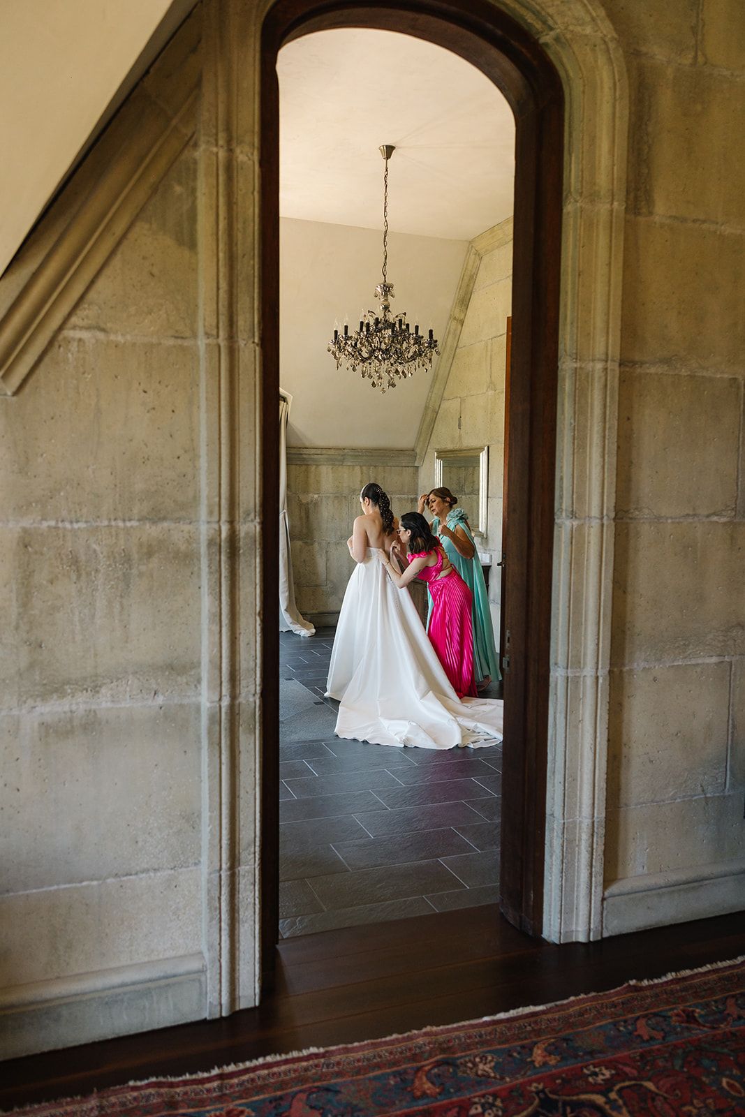 Elegant bridal party seen through an arched doorway with chandelier in a grand stone mansion.