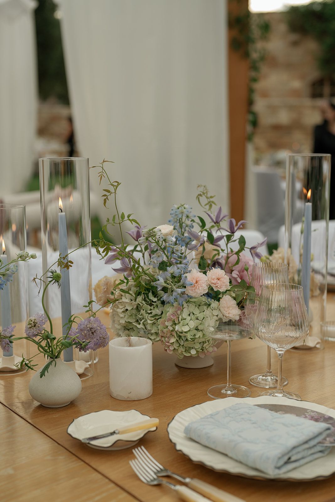 Elegant wedding table setting with pastel floral centerpiece, tall glass candles, fine china, and linen napkins on wood table.