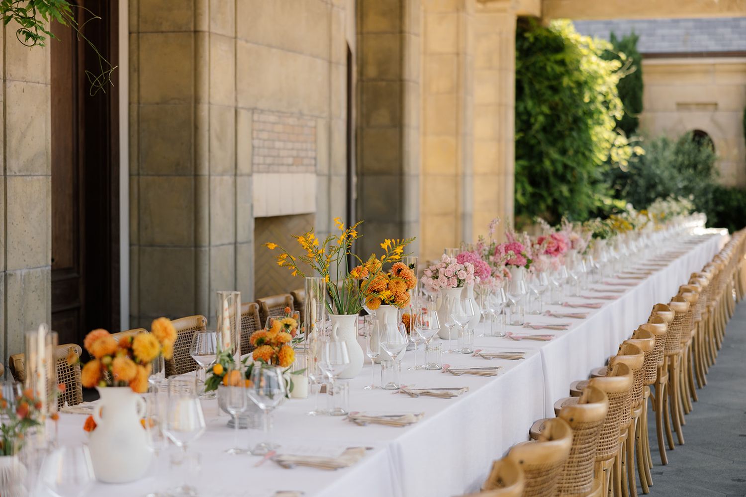Long outdoor wedding reception table set with white linens and orange floral centerpieces along a stone colonnade.