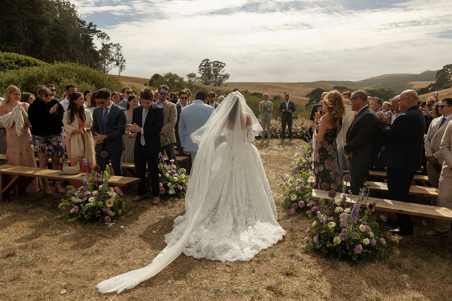 Bride in white gown and veil walks down outdoor aisle at rustic countryside wedding ceremony with floral arrangements.