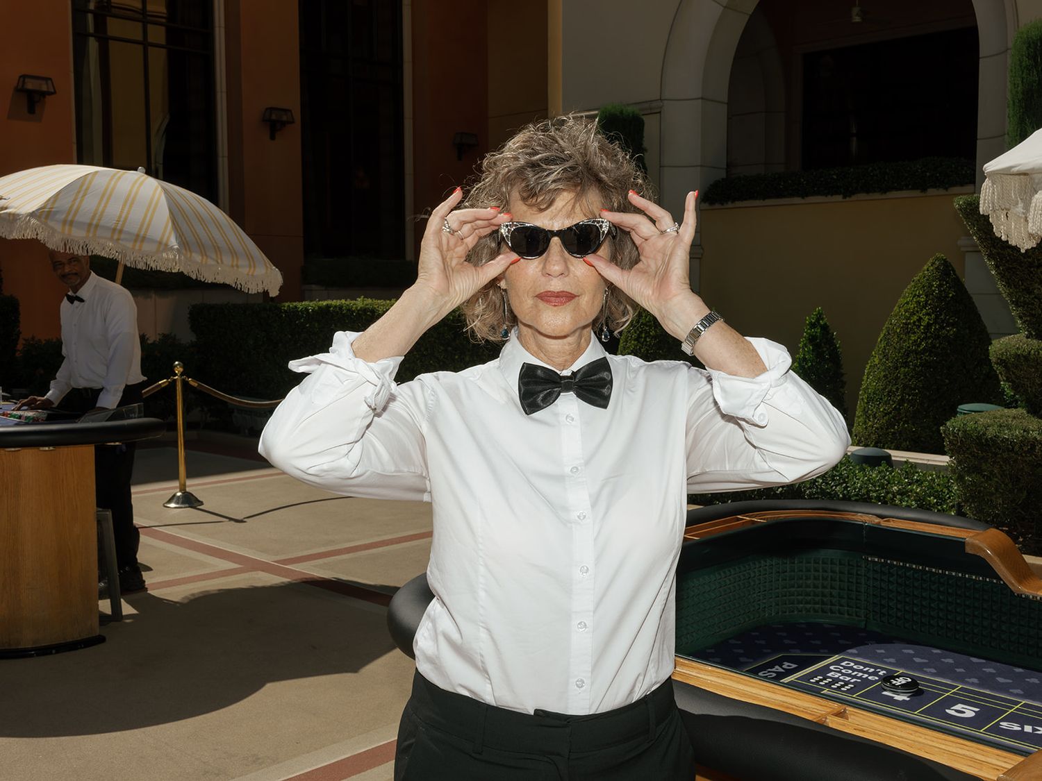 Woman in white shirt and bow tie adjusting stylish sunglasses outdoors at a casino event.
