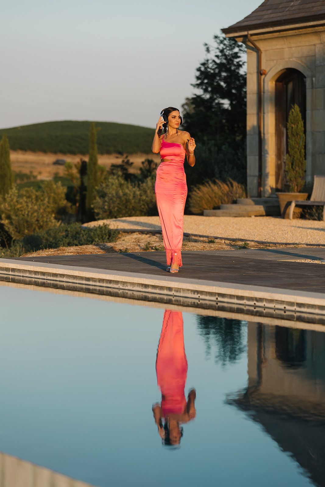 Woman in coral dress reflected in infinity pool during sunset at luxury venue.