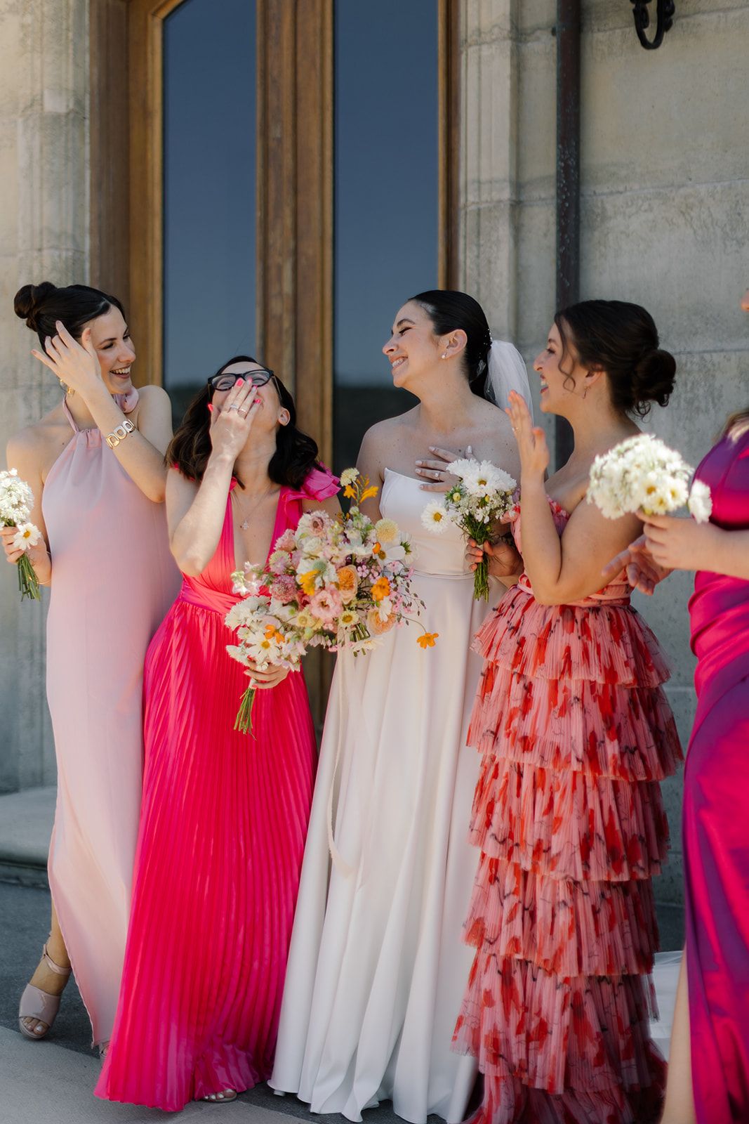 Bridesmaids in colorful pink and coral dresses laughing together while holding bouquets.