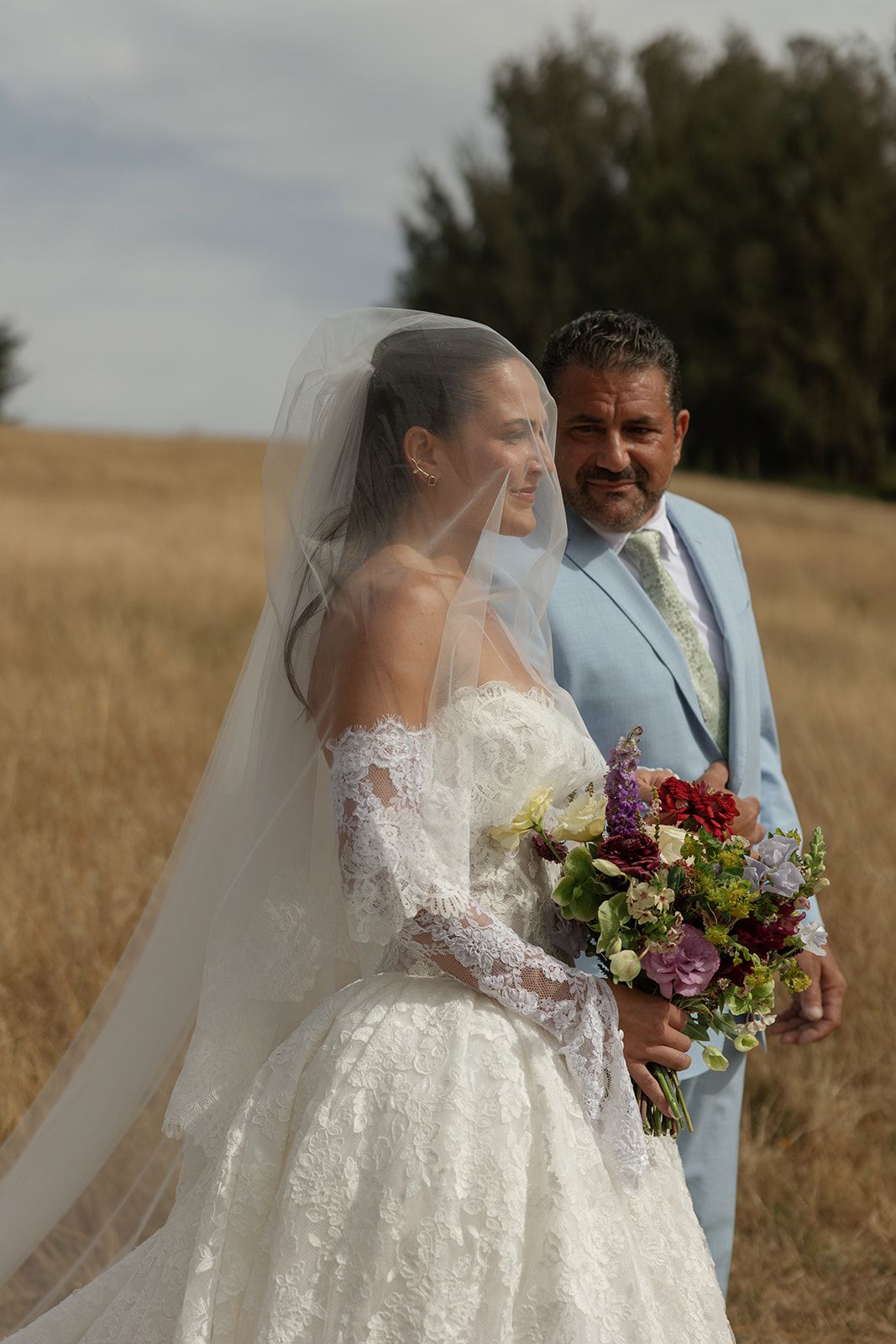 Bride in lace gown and veil holds colorful bouquet beside groom in light blue suit in golden field.