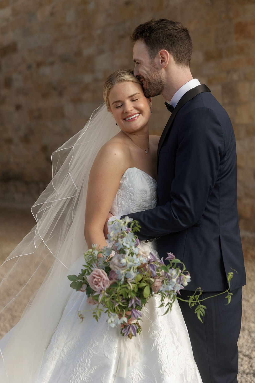 Bride in white gown with veil holds purple bouquet as groom in navy suit embraces her, smiling outdoors.