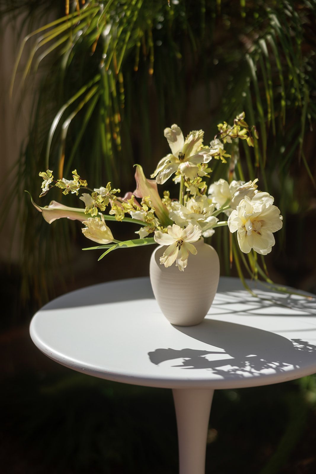 White ceramic vase with cream flowers on a round white table, set against lush green tropical foliage in sunlight.