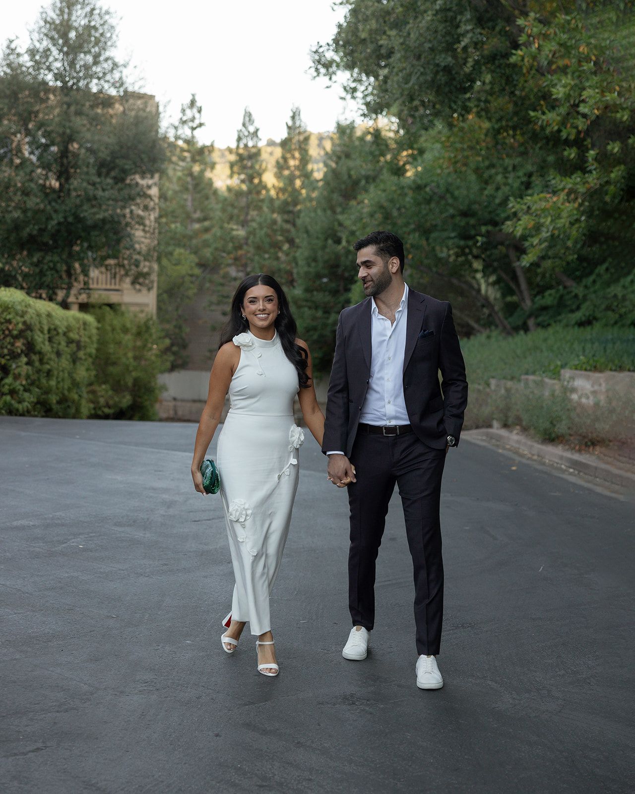 A couple walking together on a paved road surrounded by trees at dusk, wearing formal attire.