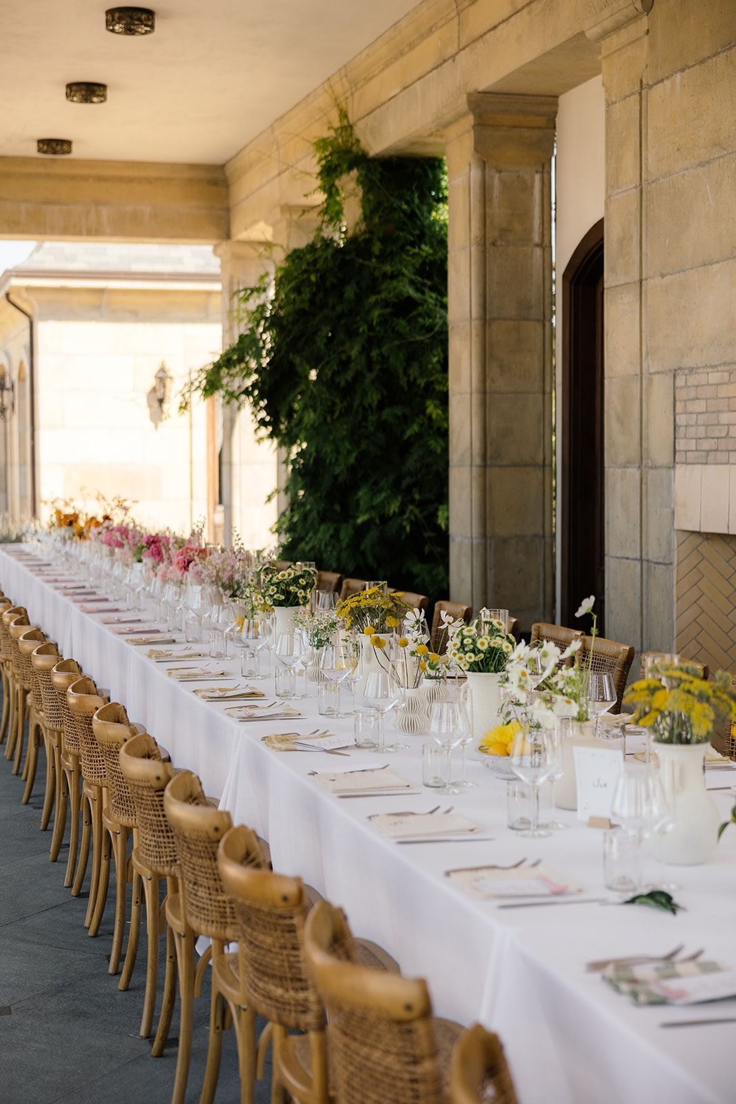 Long white dining table with rattan chairs set for elegant outdoor wedding reception.