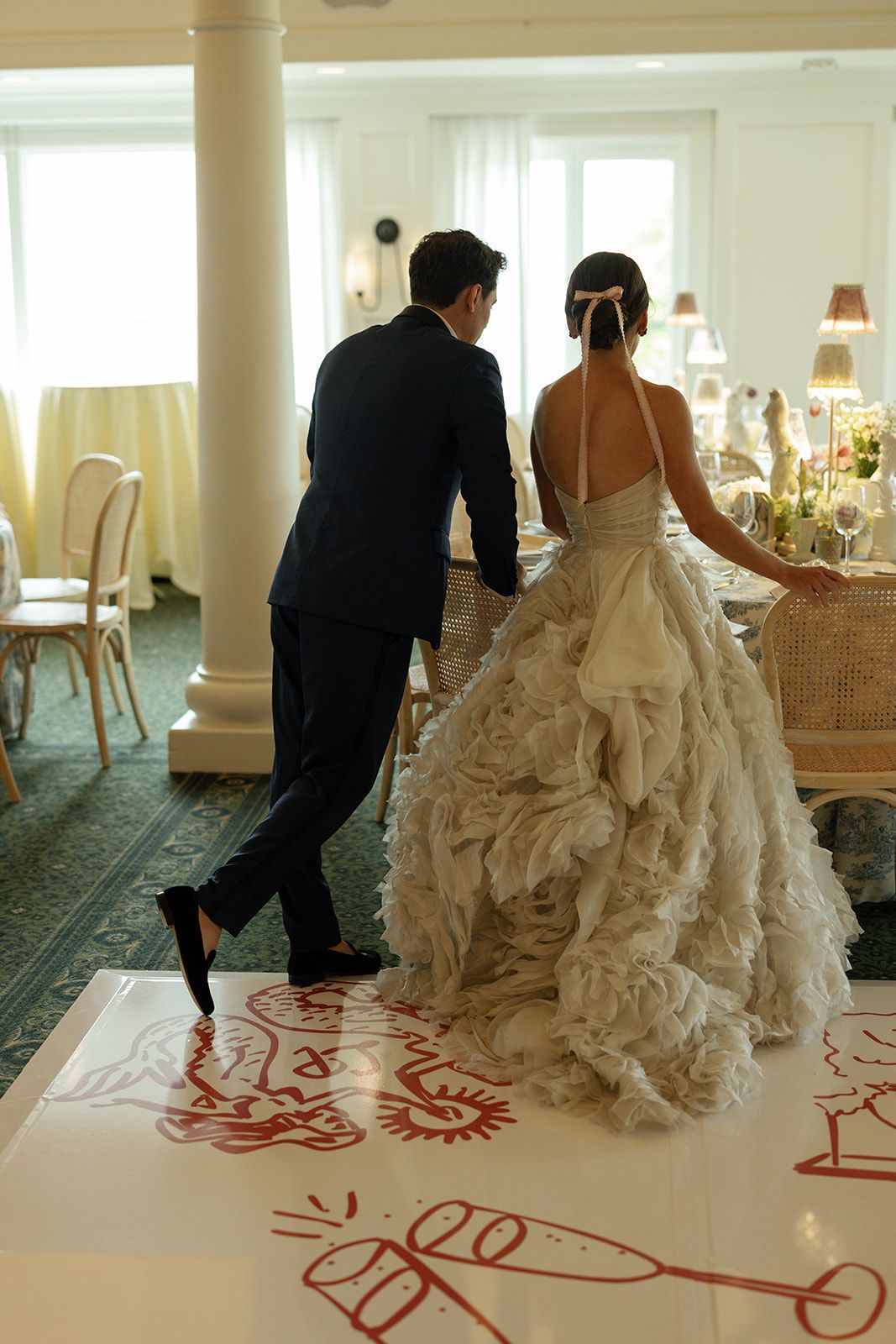 A couple in formal wedding attire stands on a patterned dance floor in an elegant room with soft lighting and decor.