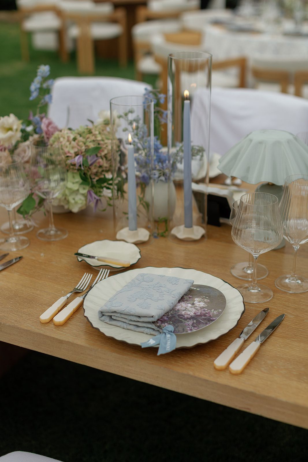 Elegant outdoor wedding table setting with blue candles, floral centerpiece, fine china, and ivory-handled cutlery on wood table.