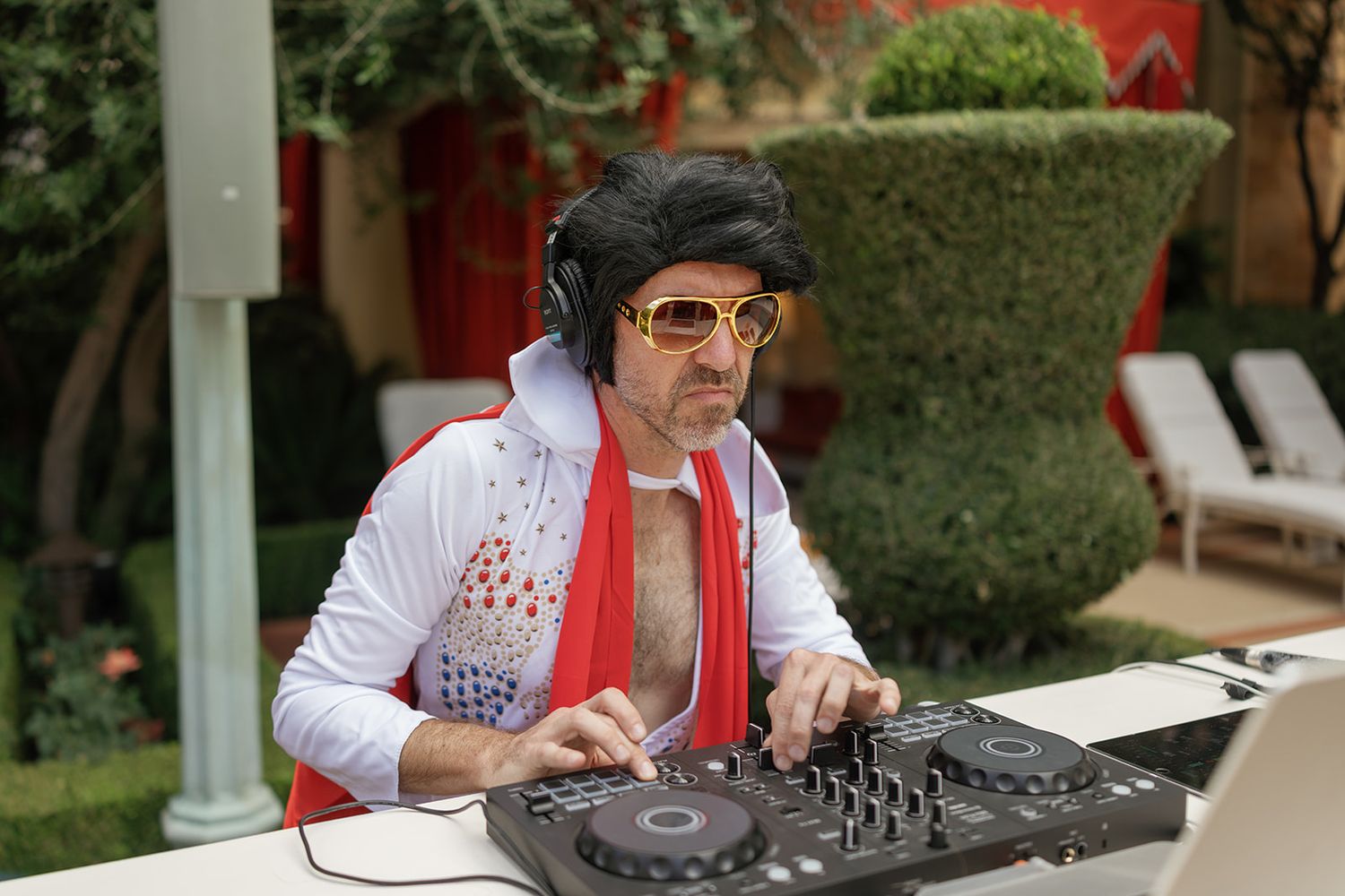 Elvis impersonator DJ in white jumpsuit, red scarf, black wig, and sunglasses mixing music at outdoor DJ booth.