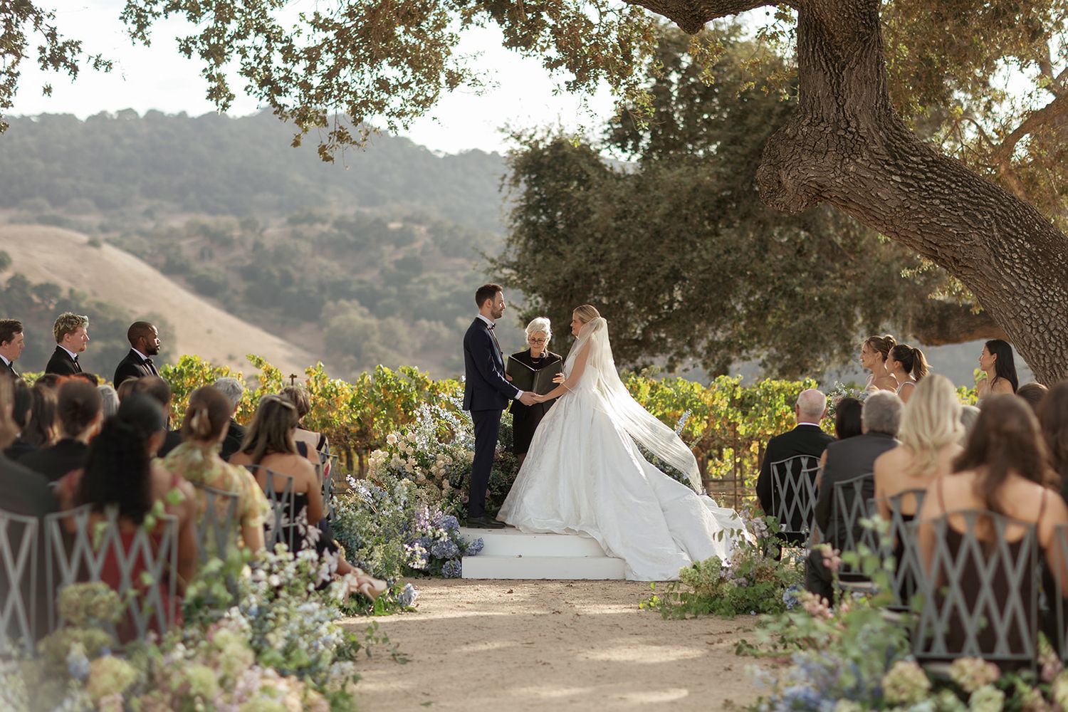 Outdoor vineyard wedding ceremony under oak tree with rolling hills, bride in white gown, guests seated along floral aisle.