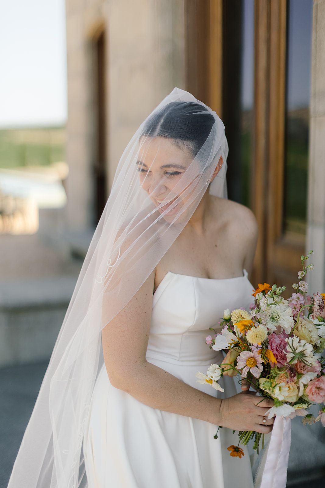 Ethereal bridal portrait with flowing veil and colorful bouquet against stone architecture.