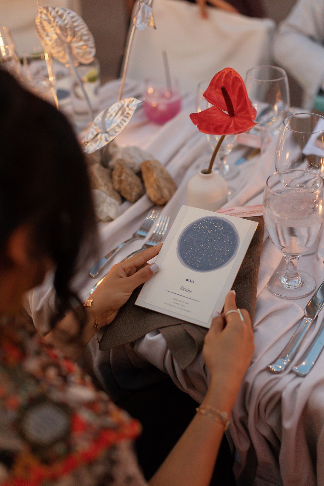 Someone reads a menu or program card with a circular design at a formal table setting with wine glasses and red flowers.