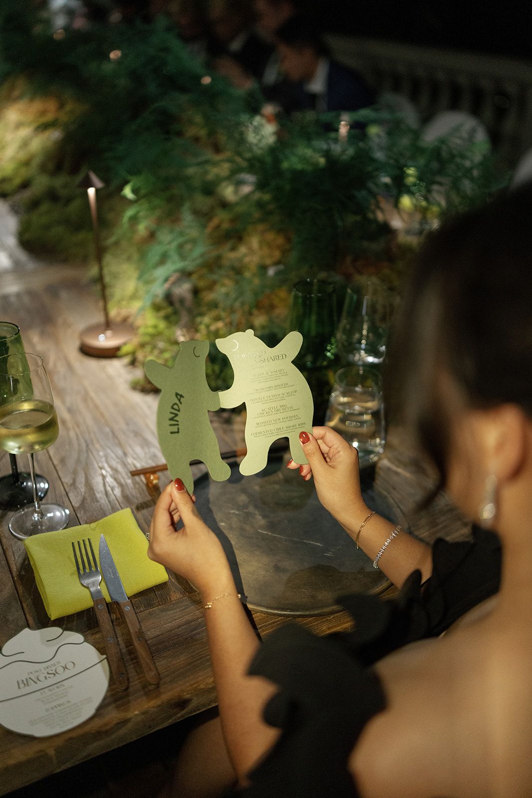 Woman holding a bear-shaped menu card at an elegant dinner event with greenery and candlelight.