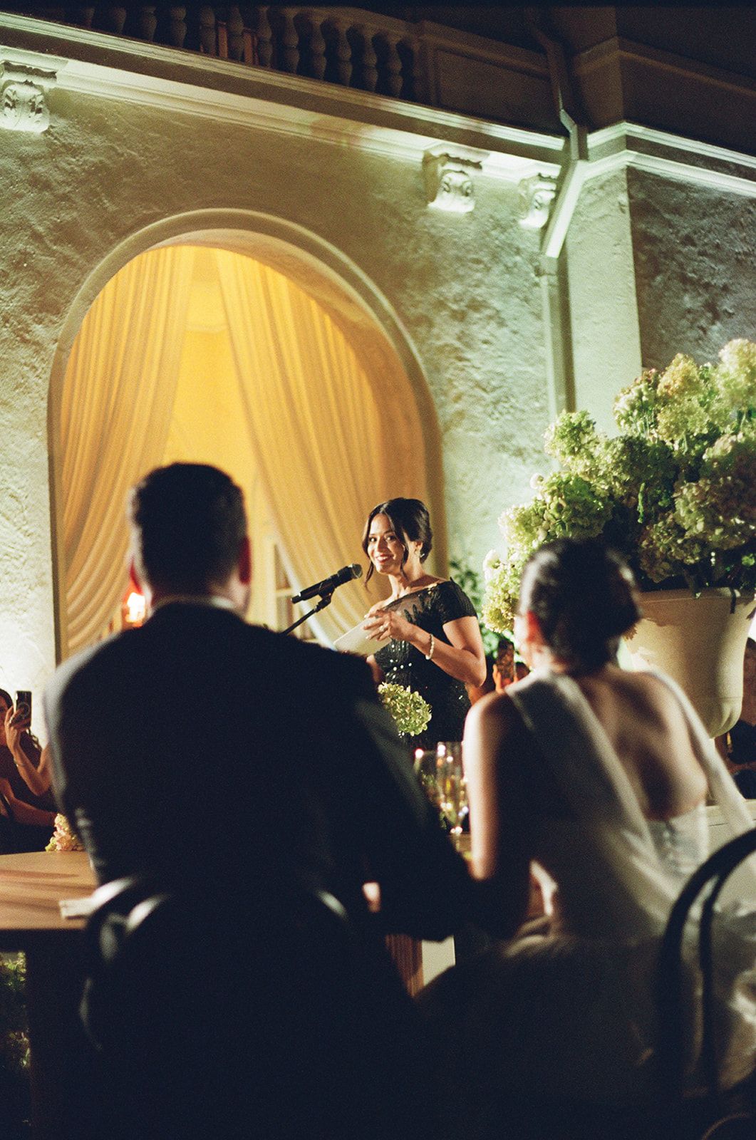 Evening reception speech in elegant archway setting with warm lighting and floral arrangements.