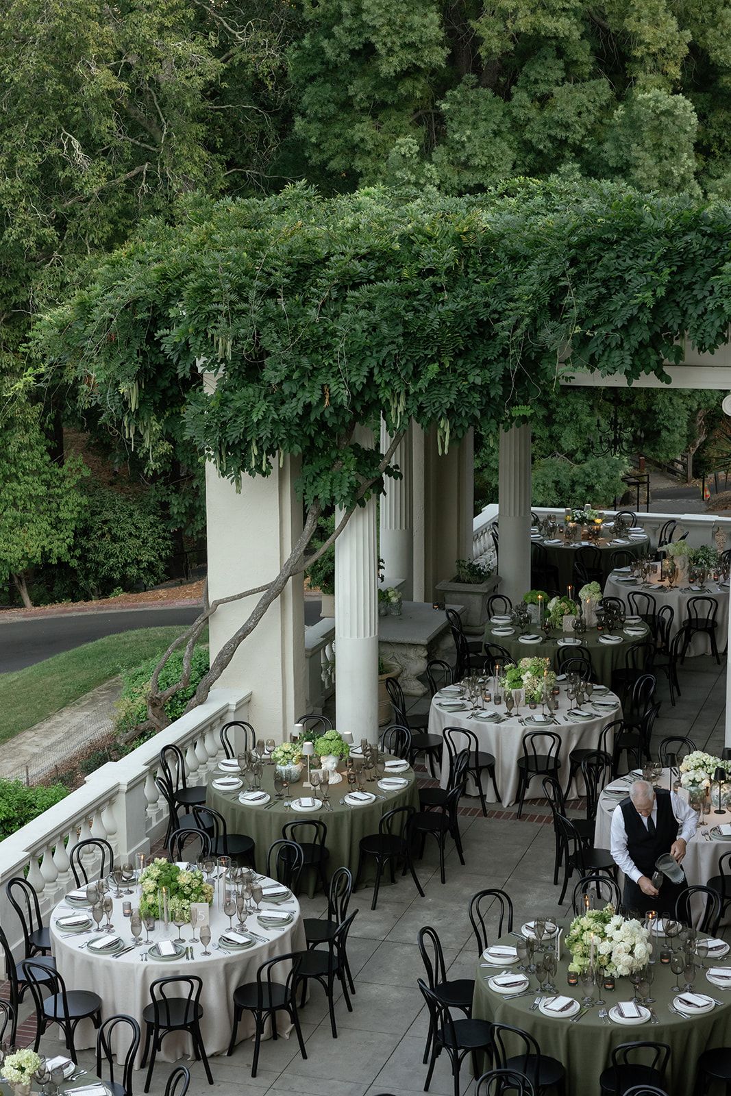 Elegant outdoor wedding reception setup with round tables, black chairs, and lush greenery under a covered patio area.
