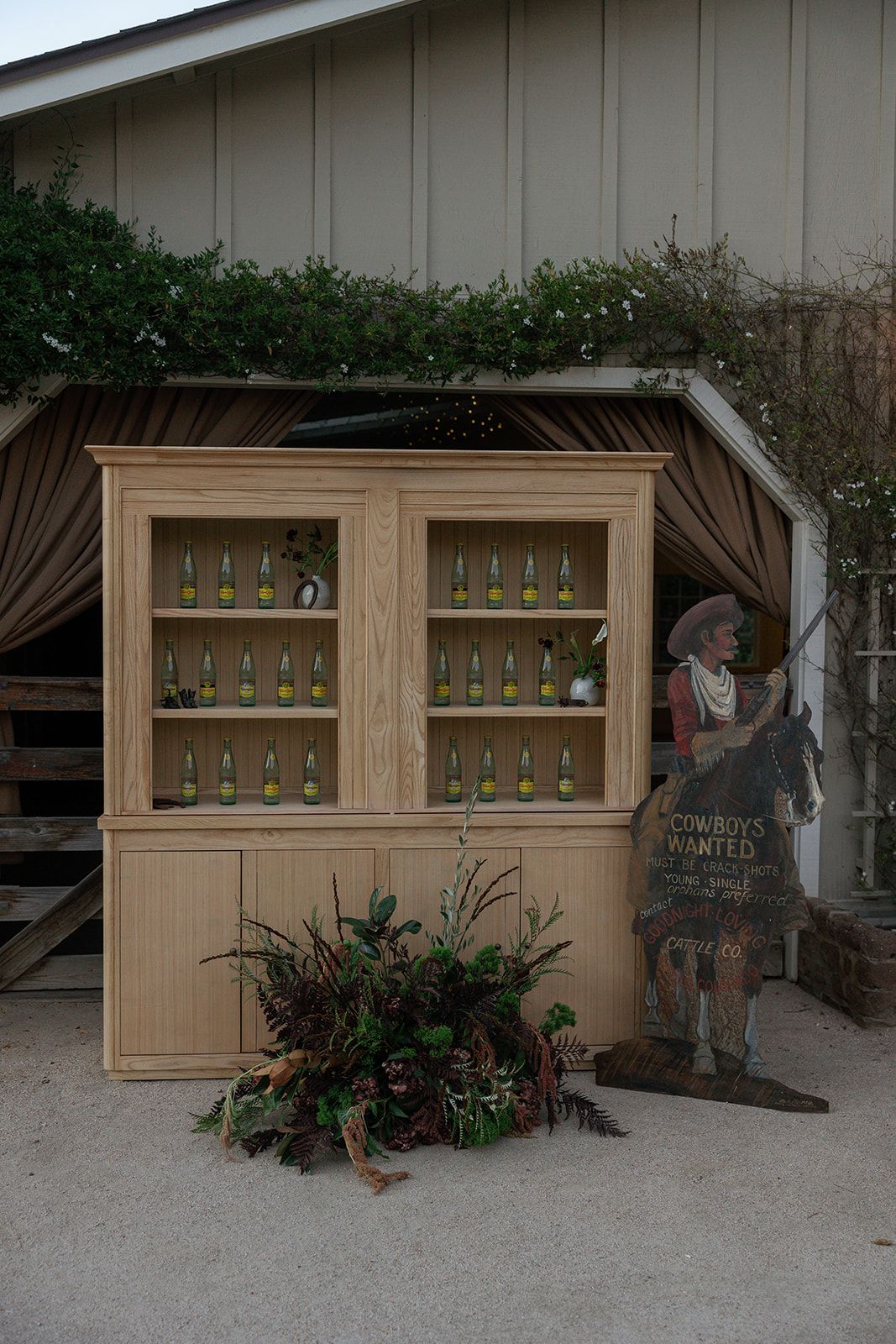 Rustic wooden display cabinet with wine bottles and decorative metal figure stands against beige wall outdoors.