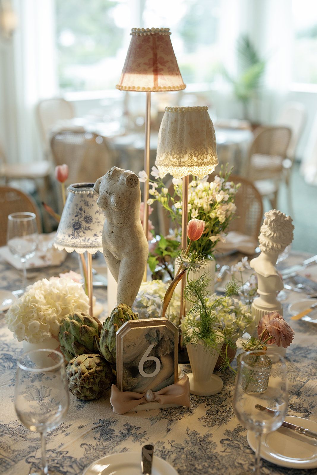 Elegant wedding table centerpiece with vintage lamps, lace shades, white flowers and a cherub statue on blue toile tablecloth.