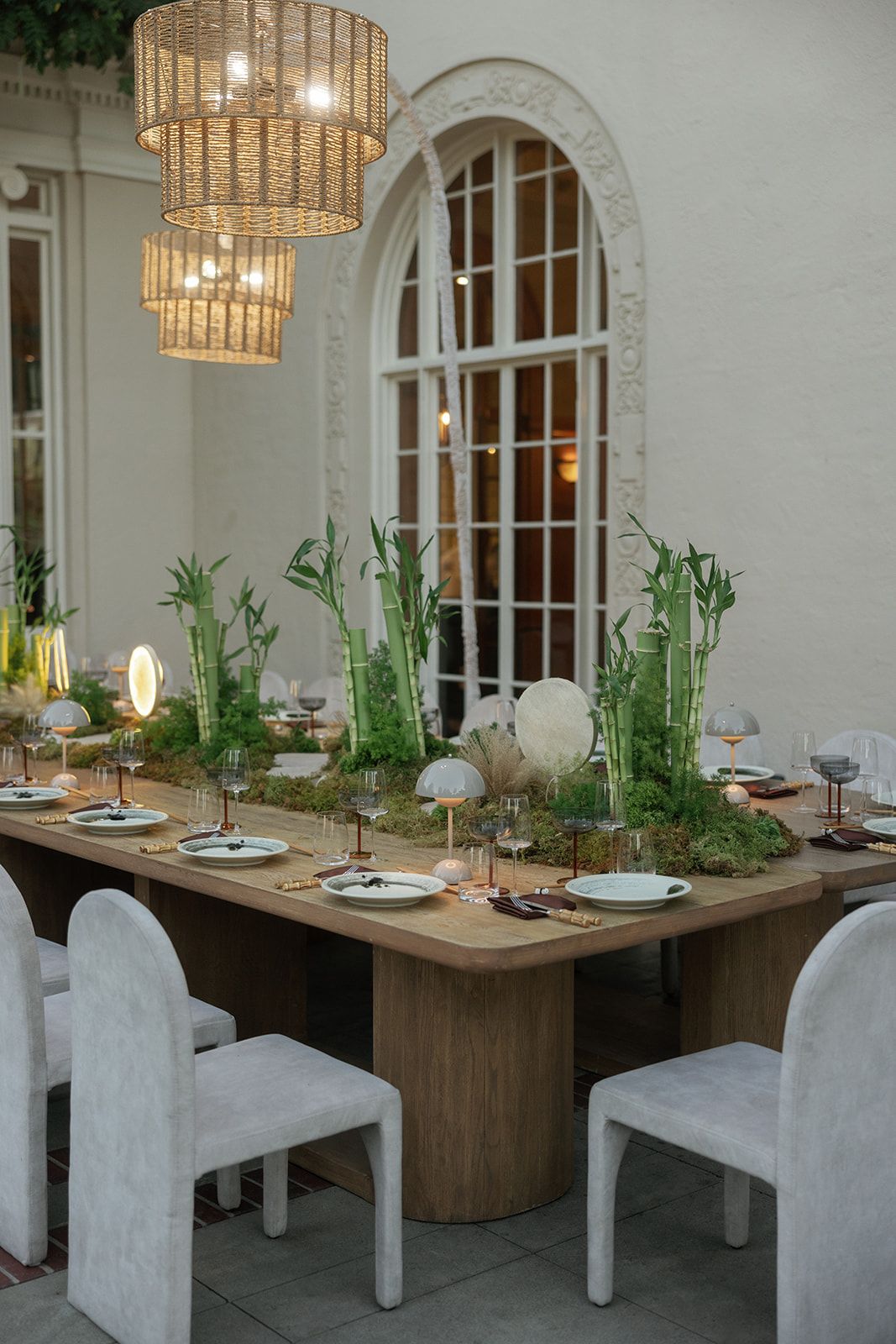 Elegant dining table with bamboo centerpiece, mushroom lamps, white chairs, and wicker chandeliers near an arched window.