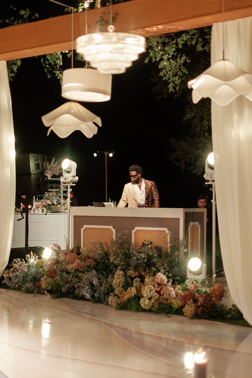 DJ booth decorated with pastel floral arrangements under white pendant lights at elegant outdoor wedding reception.