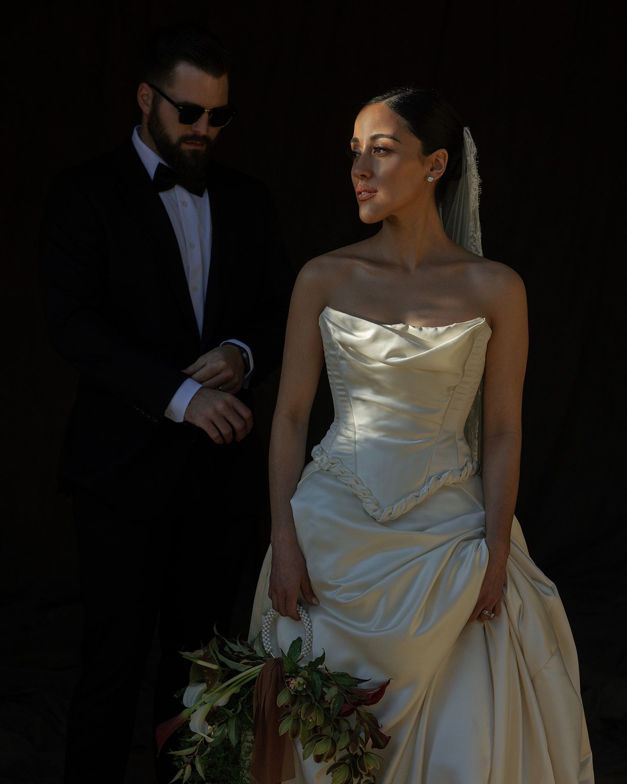 A couple in formal wedding attire poses together against a dark background with dramatic lighting.