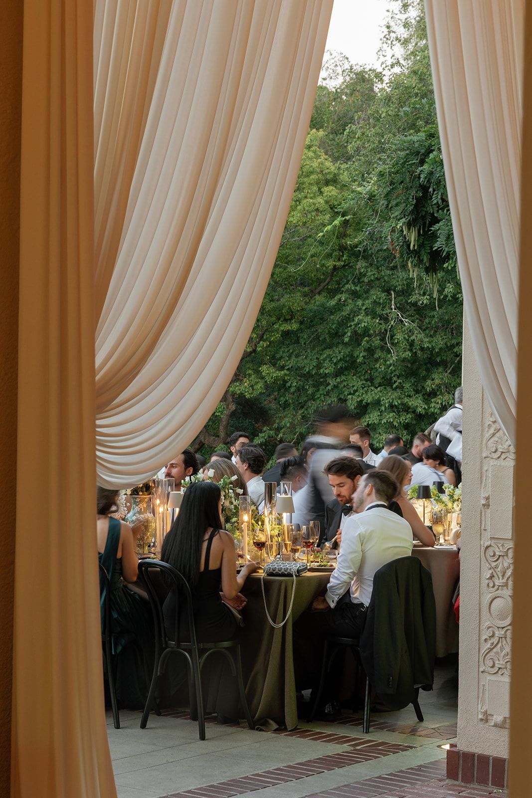 Elegant wedding reception dinner setup with flowing cream curtains and guests seated at tables under outdoor lighting.