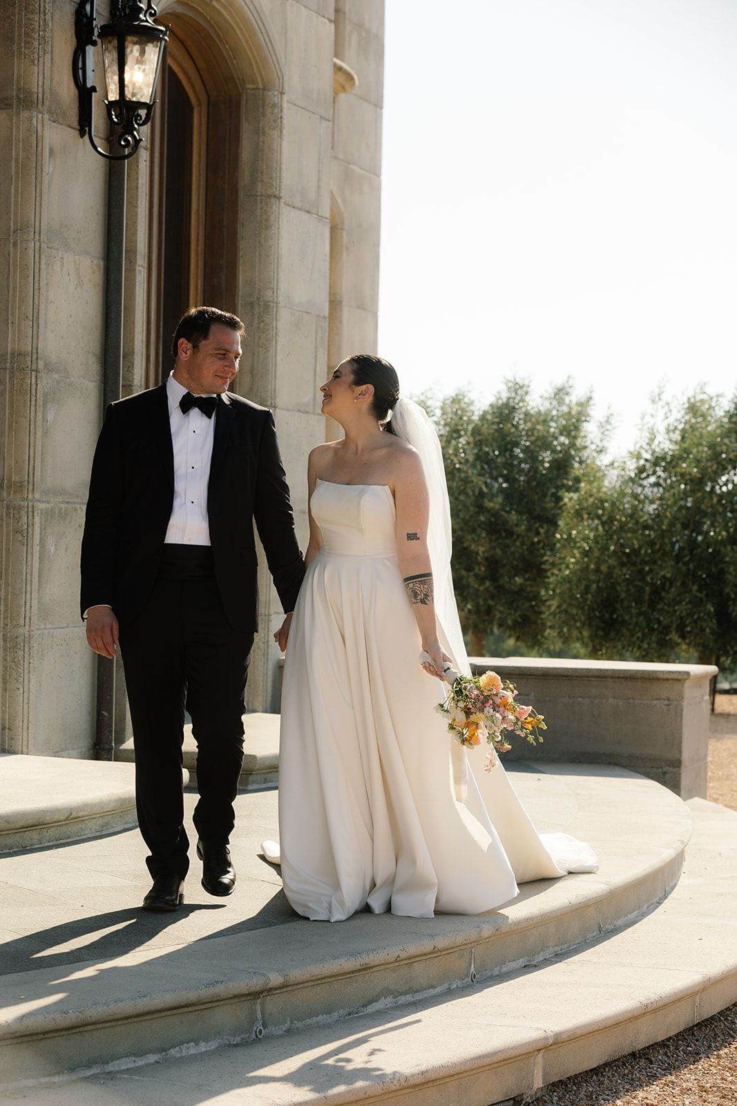 Elegant wedding couple poses on stone terrace with classic architecture and greenery backdrop.