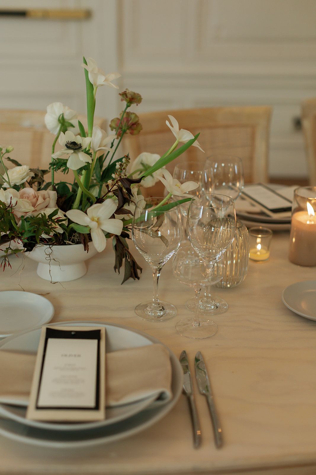 Elegant table setting with white floral centerpieces, crystal wine glasses, and candlelight on a light wooden table.