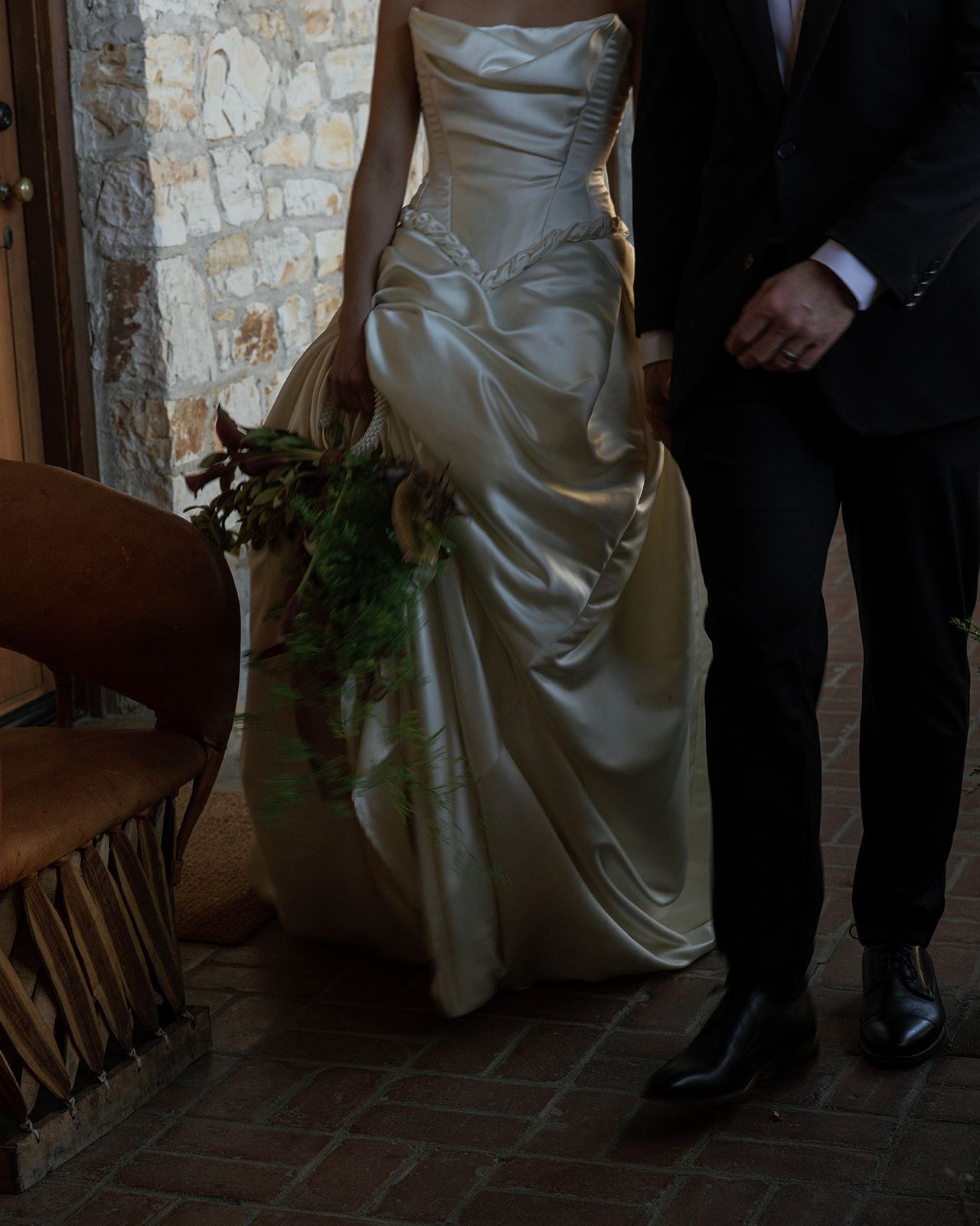 Elegant white satin wedding dress with ruched detailing shown next to formal black suit in rustic stone setting.