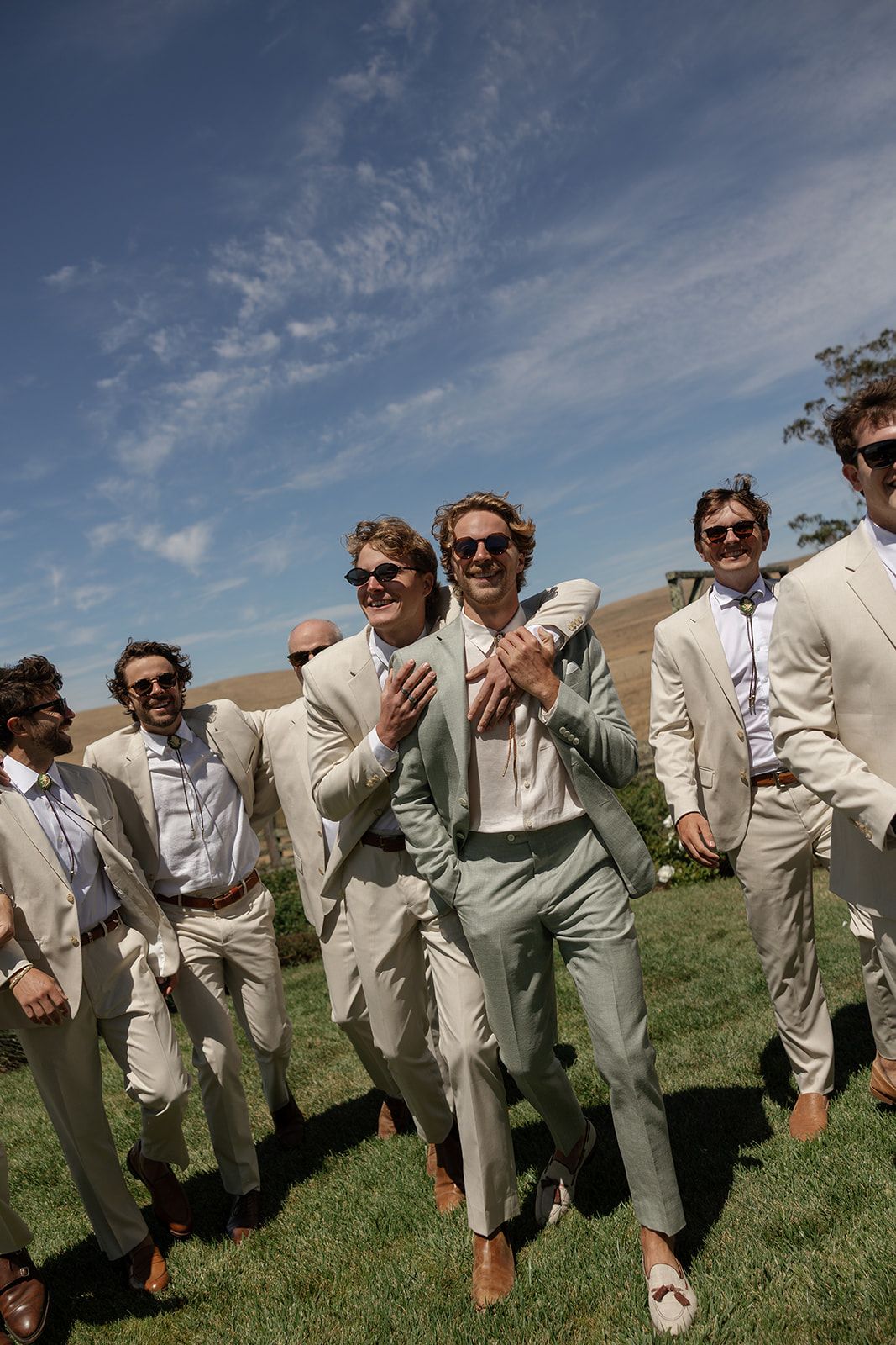 Groomsmen in tan suits walk outdoors on green grass under a blue sky, with the groom in a mint suit clapping.