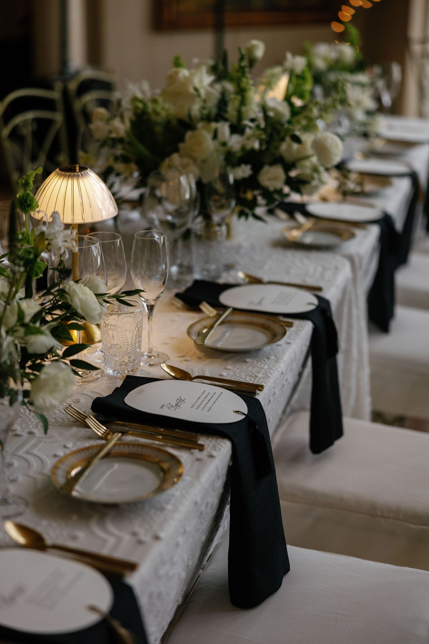 Elegant table settings with white plates, gold cutlery, black napkins, and greenery decorations at a formal dinner event.