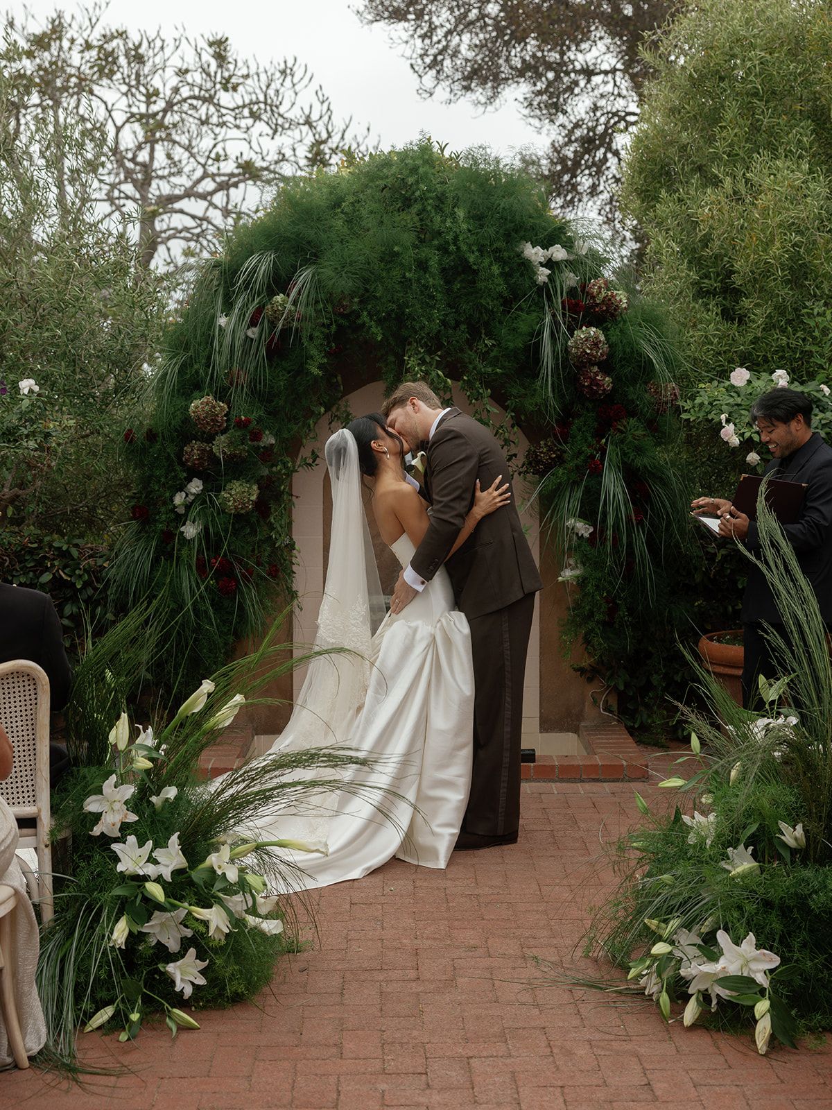 Bride and groom share first kiss under lush green floral arch with white lilies at outdoor garden wedding ceremony.