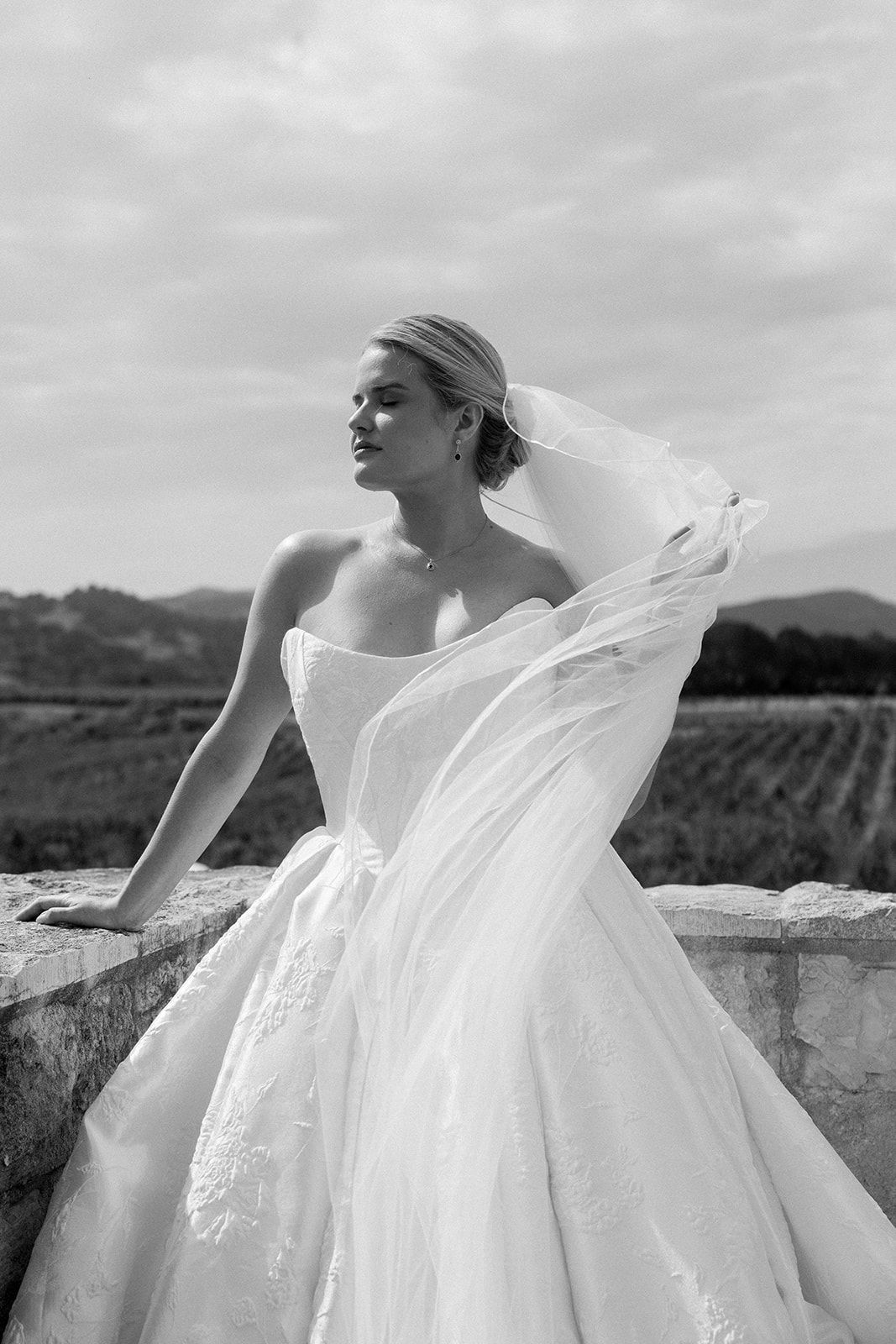 Black and white photo of a bride in a strapless ballgown with flowing veil outdoors.