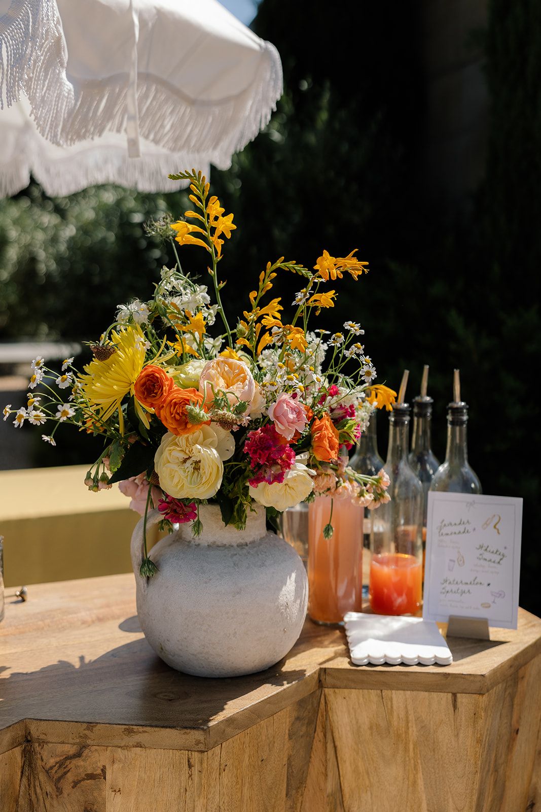 Colorful floral arrangement in white vase with cocktail bottles and menu card on rustic wooden display.