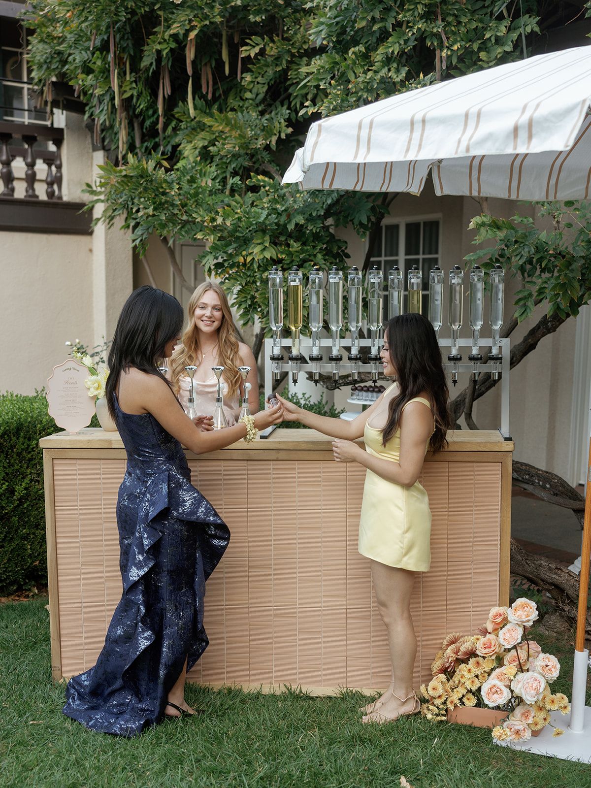 Three women in elegant dresses gather around an outdoor bar decorated with flowers at a garden party.